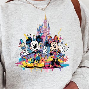 Peut inclure: Sweat-shirt gris clair avec un motif color&eacute; de Mickey et Minnie Mouse devant un ch&acirc;teau. Mickey tient un pinceau, Minnie fait le signe de la paix, et tous deux sont entour&eacute;s d'&eacute;claboussures de peinture et de notes de musique.