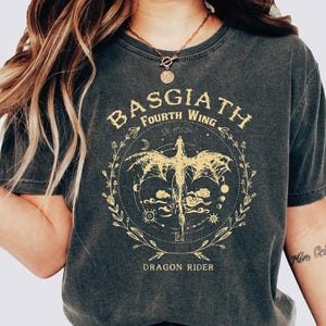 May include: Dark gray t-shirt featuring a gold dragon design within a circular frame. The text "BASGIATH FOURTH WING" arches above the dragon, with "DRAGON RIDER" below. The design includes celestial elements and floral accents.