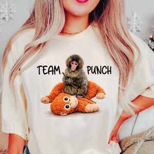 May include: Cream-colored t-shirt with the text "TEAM PUNCH" and a graphic of two monkeys. One monkey is sitting on top of the other, holding a stick. The bottom monkey is orange, and the top monkey is gray.
