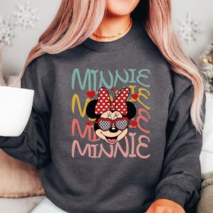 Puede incluir: Sudadera gris oscuro con un dise&ntilde;o de Minnie Mouse. El dise&ntilde;o incluye la palabra "Minnie" en varios colores, una cara de Minnie Mouse con gafas de sol en forma de coraz&oacute;n y un lazo rojo con lunares blancos. Cuello redondo.