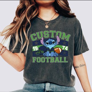 May include: Dark grey t-shirt with a cartoon Stitch character in a football uniform. The shirt displays the words "CUSTOM FOOTBALL" in green and white, with the numbers "19" and "74". The design includes a football and helmet.