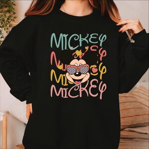May include: Black sweatshirt featuring the word "MICKEY" in colorful, layered text. The design includes a cartoon Mickey Mouse face wearing heart-shaped sunglasses. The overall design is playful and vibrant.