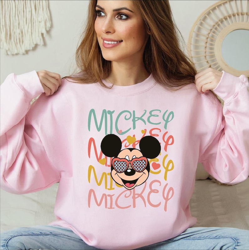 May include: A light pink sweatshirt featuring the word "Mickey" in multiple colors and a graphic of Mickey Mouse wearing heart-shaped sunglasses. The sweatshirt is a crew neck style.