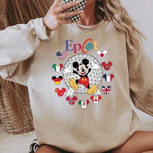 May include: Beige sweatshirt featuring the word "Epcot" in colorful letters, with a globe graphic and Mickey Mouse. Mickey is surrounded by Mickey Mouse-shaped flags representing different countries. The sweatshirt has long sleeves.