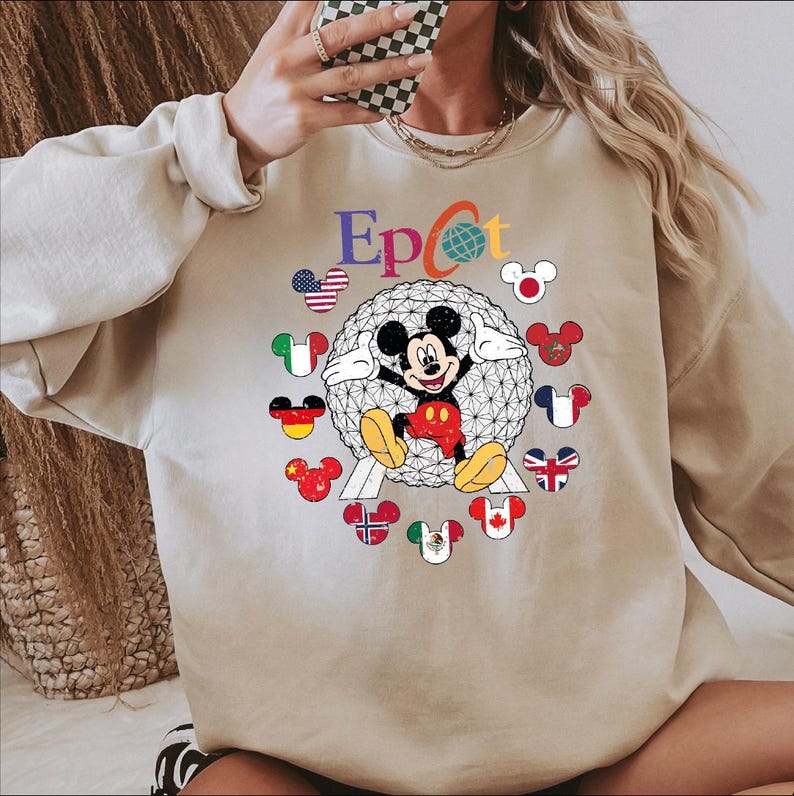 May include: Beige sweatshirt featuring the word "Epcot" in colorful letters, with a globe graphic and Mickey Mouse. Mickey is surrounded by Mickey Mouse-shaped flags representing different countries. The sweatshirt has long sleeves.