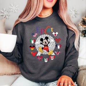 May include: Dark gray sweatshirt featuring a colorful "Epcot" design with Mickey Mouse and various country flags. The design includes a geometric sphere and Mickey Mouse ears with flags around the sphere.