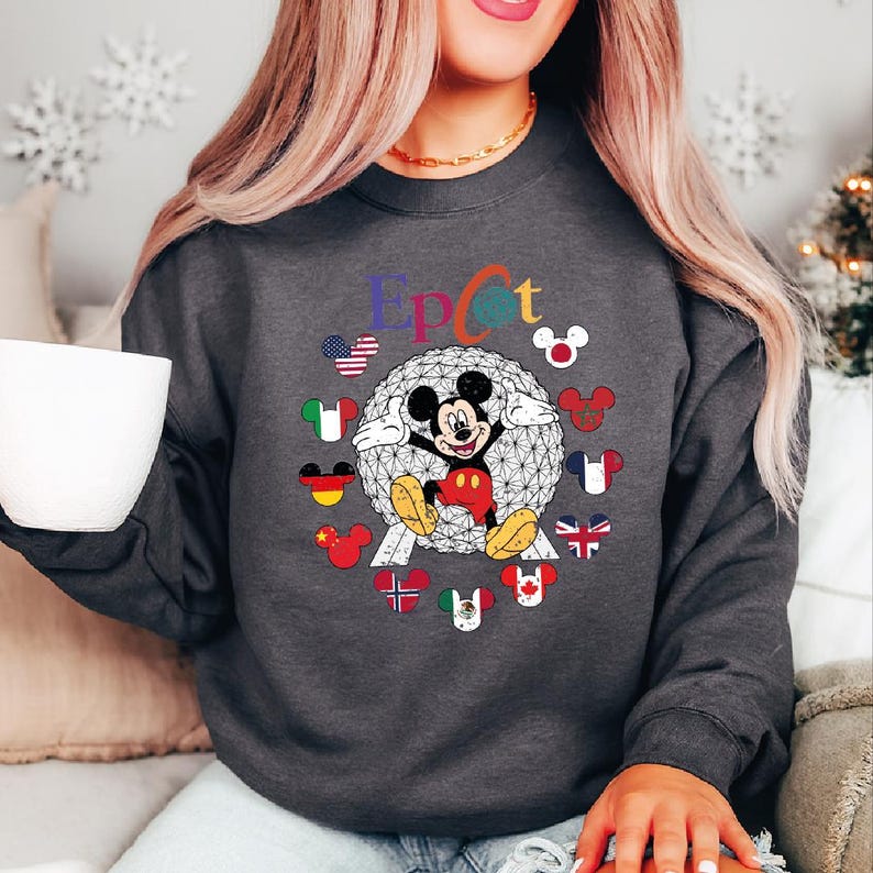 May include: Dark gray sweatshirt featuring a colorful "Epcot" design with Mickey Mouse and various country flags. The design includes a geometric sphere and Mickey Mouse ears with flags around the sphere.