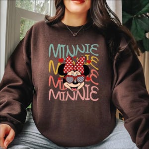 Puede incluir: Una sudadera marr&oacute;n con un gr&aacute;fico de Minnie Mouse con gafas de sol en forma de coraz&oacute;n y un lazo de lunares. La palabra "MINNIE" est&aacute; apilada verticalmente en letras coloridas. Un dise&ntilde;o divertido y juguet&oacute;n.
