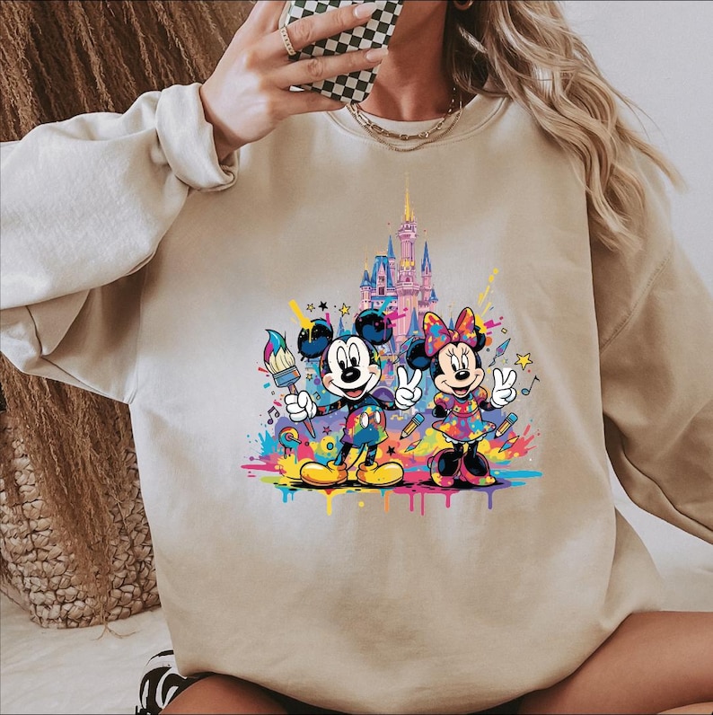 May include: Beige sweatshirt featuring a colorful graphic of Mickey and Minnie Mouse with a castle backdrop. Mickey holds a paintbrush, Minnie makes a peace sign, and both are surrounded by paint splatters and stars.