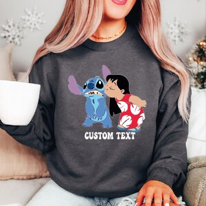Pode incluir: Camisola cinza escuro com um gr&aacute;fico de Stitch e Lilo a beijarem-se. Stitch &eacute; azul com orelhas grandes, e Lilo usa um vestido vermelho com padr&otilde;es florais brancos. O texto "CUSTOM TEXT" est&aacute; impresso abaixo.