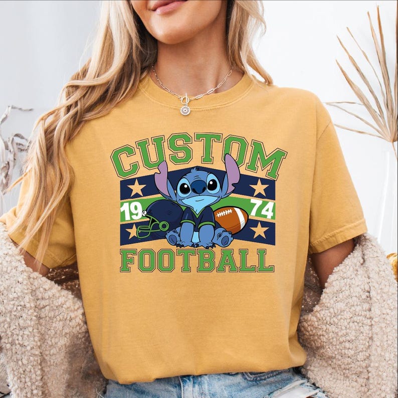 May include: Mustard yellow t-shirt with a blue cartoon character holding a football. The shirt has the words "CUSTOM FOOTBALL" in green and white, with the numbers "19" and "74" on the design. The character is wearing a football uniform.