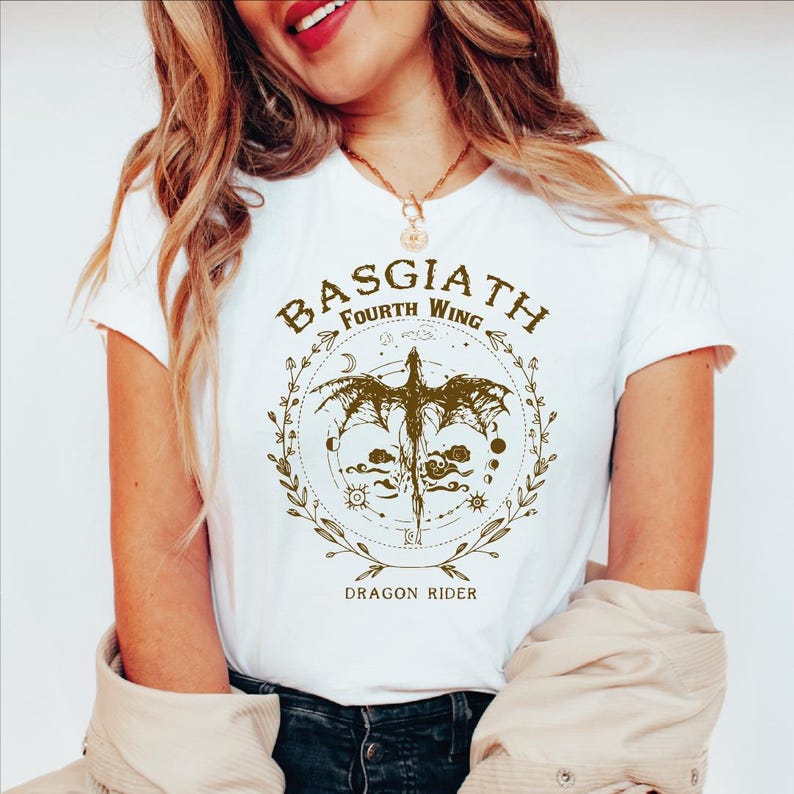 May include: White t-shirt featuring a detailed graphic with the text "BASGIATH Fourth Wing" and "DRAGON RIDER". The design includes a dragon, celestial elements, and decorative borders in a gold color. The shirt has short sleeves.