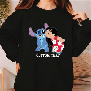 Pode incluir: Camisola preta com um gr&aacute;fico de Stitch e Lilo de Lilo & Stitch da Disney. Lilo beija Stitch na bochecha. As palavras "CUSTOM TEXT" s&atilde;o impressas em branco. A camisola &eacute; de gola redonda.