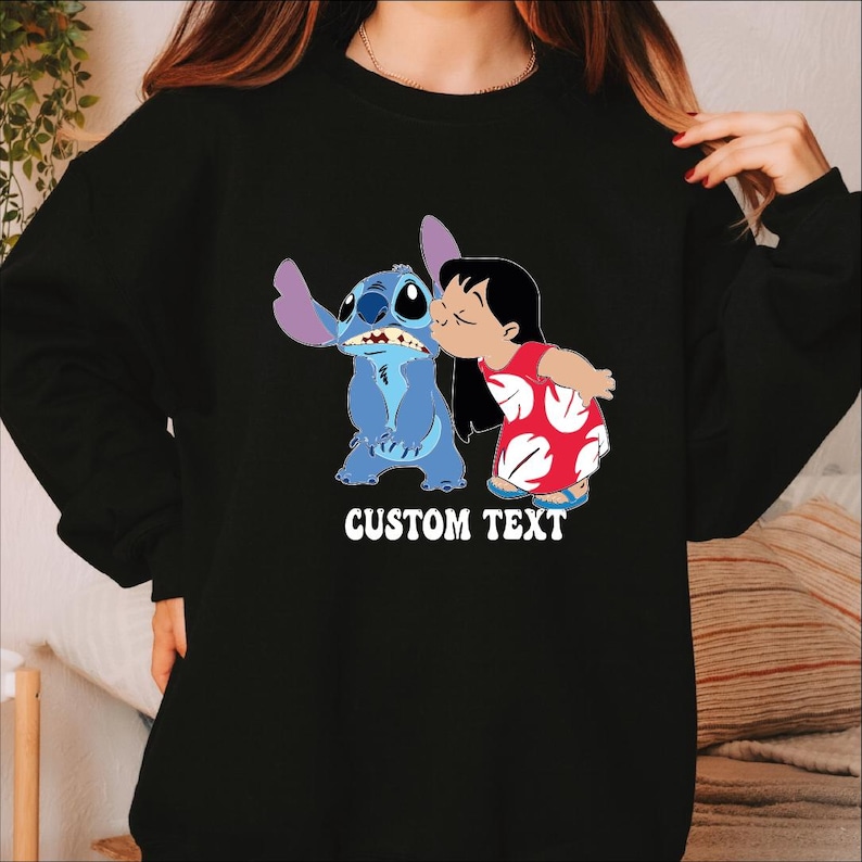 Pode incluir: Camisola preta com um gr&aacute;fico de Stitch e Lilo de Lilo & Stitch da Disney. Lilo beija Stitch na bochecha. As palavras "CUSTOM TEXT" s&atilde;o impressas em branco. A camisola &eacute; de gola redonda.