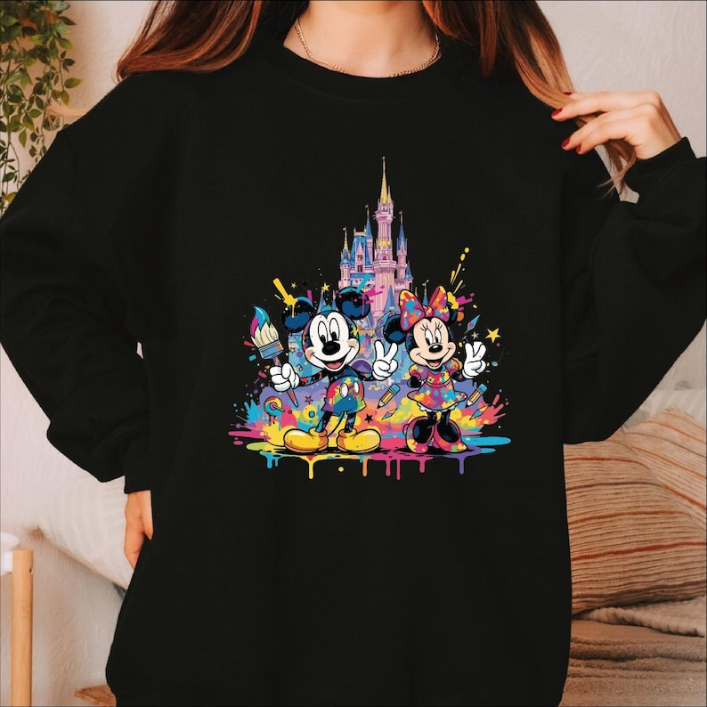 Peut inclure: Sweat-shirt noir avec Mickey et Minnie Mouse dans un style color&eacute;. Le motif comprend un ch&acirc;teau et les personnages tenant des pinceaux et des crayons, avec des &eacute;claboussures de peinture vives en jaune, bleu, rose et vert.