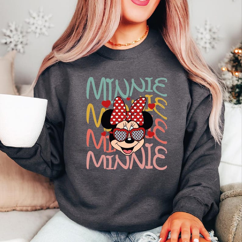 Puede incluir: Sudadera gris oscuro con un dise&ntilde;o de Minnie Mouse. El dise&ntilde;o incluye la palabra "Minnie" en varios colores, una cara de Minnie Mouse con gafas de sol en forma de coraz&oacute;n y un lazo rojo con lunares blancos. Cuello redondo.