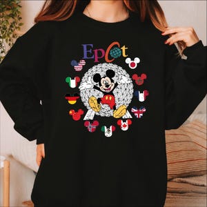 May include: Black sweatshirt featuring the word "Epcot" in colorful letters. The design includes a geometric sphere with Mickey Mouse in the center, surrounded by flags and Mickey Mouse icons representing different countries. A fun and colorful design.