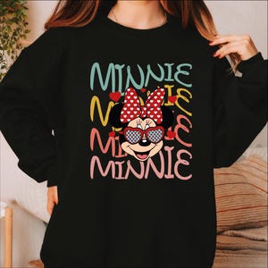 Puede incluir: Sudadera negra con un dise&ntilde;o de Minnie Mouse. La palabra "MINNIE" est&aacute; apilada verticalmente en varios colores, con Minnie Mouse usando gafas de sol en forma de coraz&oacute;n y un lazo de lunares. El dise&ntilde;o tiene una est&eacute;tica juguetona y de dibujos animados.