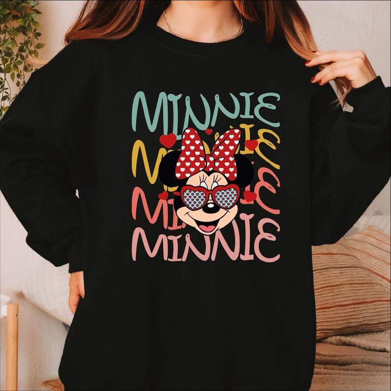 Puede incluir: Sudadera negra con un dise&ntilde;o de Minnie Mouse. La palabra "MINNIE" est&aacute; apilada verticalmente en varios colores, con Minnie Mouse usando gafas de sol en forma de coraz&oacute;n y un lazo de lunares. El dise&ntilde;o tiene una est&eacute;tica juguetona y de dibujos animados.