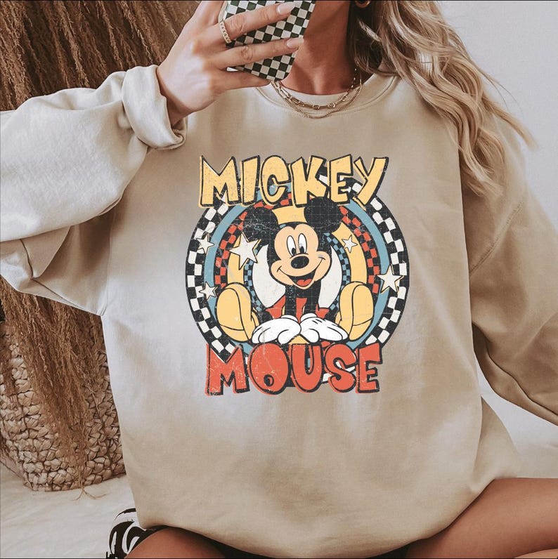 K&ouml;nnte beinhalten: Beiges Sweatshirt mit einem Retro-Mickey-Mouse-Motiv. Das Design zeigt Mickey Mouse und die Worte "Mickey Mouse" in einer Vintage-Schrift vor einem farbenfrohen kreisf&ouml;rmigen Hintergrund mit Sternen und einem Schachbrettmuster.