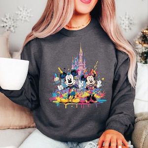 Peut inclure: Sweat-shirt gris fonc&eacute; avec un motif color&eacute; de Mickey et Minnie Mouse devant un ch&acirc;teau. Le design comprend des &eacute;claboussures de peinture et des fournitures artistiques, cr&eacute;ant une esth&eacute;tique vibrante et ludique. Le sweat-shirt est de style d&eacute;contract&eacute; et confortable.