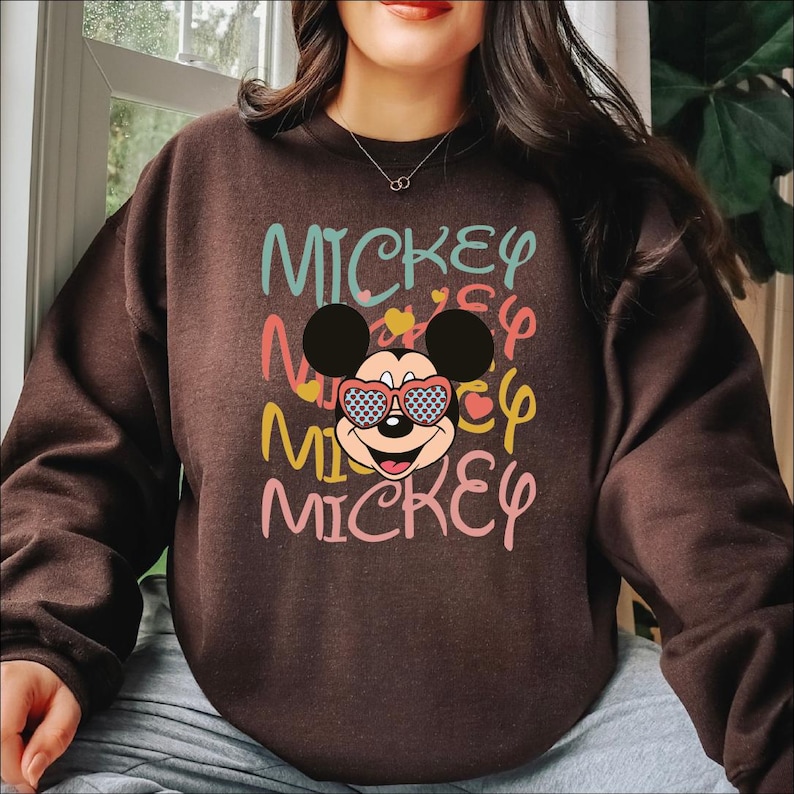 May include: A brown sweatshirt featuring the word "Mickey" in multiple colors and a graphic of Mickey Mouse wearing heart-shaped sunglasses. The design includes hearts and a playful font.