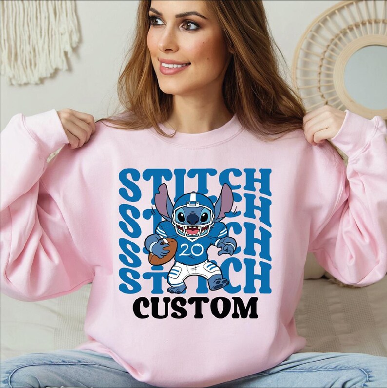 May include: A light pink sweatshirt featuring a blue graphic of Stitch in a football uniform, holding a football. The word "STITCH" is repeated in blue, with "CUSTOM" in black below the graphic.