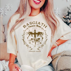 May include: Cream-colored t-shirt with a brown graphic featuring a dragon, the text "BASGIATH FOURTH WING", and "DRAGON RIDER". The design is encircled by a wreath. The shirt has short sleeves.