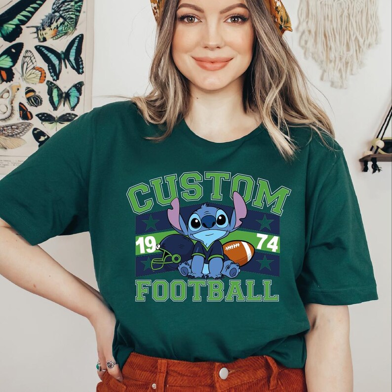 May include: Dark green t-shirt with a cartoon character holding a football. The shirt features the word "CUSTOM" above the character, with "19" and "74" on either side. The word "FOOTBALL" is printed below the character in green.