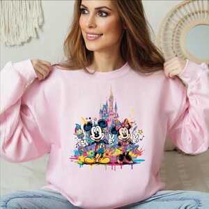 Peut inclure: Un sweat-shirt rose clair avec un motif color&eacute; de Mickey et Minnie Mouse devant un ch&acirc;teau. Mickey tient un pinceau, Minnie fait le signe de la paix. Le motif comprend des &eacute;claboussures de peinture color&eacute;es.