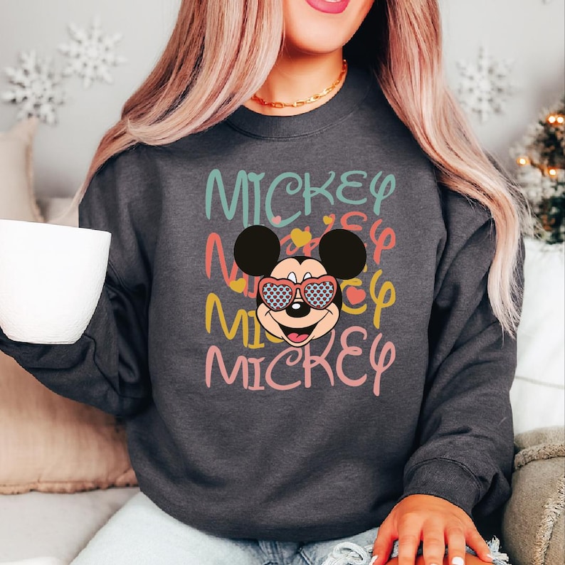 May include: Dark gray sweatshirt featuring a colorful graphic of Mickey Mouse wearing sunglasses. The word "Mickey" is repeated in various colors above and below the character. The sweatshirt is a casual style.