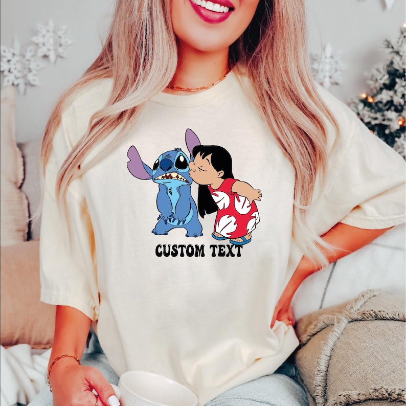 May include: Cream-colored t-shirt featuring a cartoon image of Stitch and Lilo. Lilo is kissing Stitch on the cheek. The text "CUSTOM TEXT" is printed below the image in a bold, black font. The shirt has short sleeves.