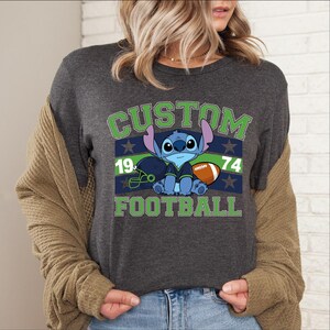 May include: Dark grey crew neck t-shirt with a cartoon character and the words "CUSTOM FOOTBALL 1974". The design features a football, helmet, and stars in green and blue. The shirt is a crew neck.