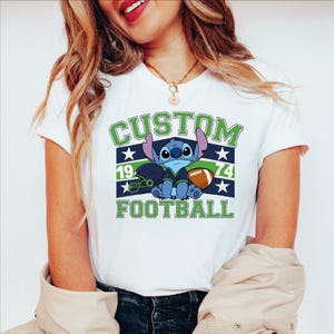 May include: White t-shirt with a cartoon character design. The design includes the text "CUSTOM FOOTBALL 1974" in green and navy blue, along with a football and stars.
