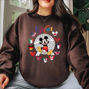 May include: A brown sweatshirt featuring a graphic of Mickey Mouse in front of a geometric sphere, with the word "Epcot" above. The design includes flags of various countries and Mickey Mouse silhouettes. The sweatshirt has long sleeves.
