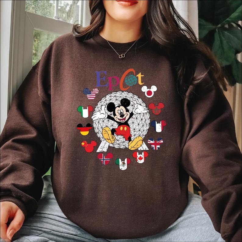 May include: A brown sweatshirt featuring a graphic of Mickey Mouse in front of a geometric sphere, with the word "Epcot" above. The design includes flags of various countries and Mickey Mouse silhouettes. The sweatshirt has long sleeves.