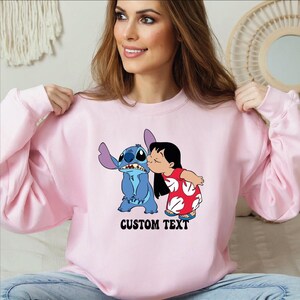 Pode incluir: Camisola rosa claro com um desenho animado de Stitch e Lilo a beijarem-se. O desenho inclui o texto "CUSTOM TEXT" em preto. A camisola tem mangas compridas.