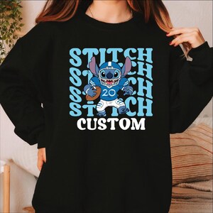 May include: Black sweatshirt featuring a cartoon Stitch character in a football uniform. The design includes the word "STITCH" repeated in light blue, with "CUSTOM" in white below the character. Stitch is wearing a blue helmet and holding a football.