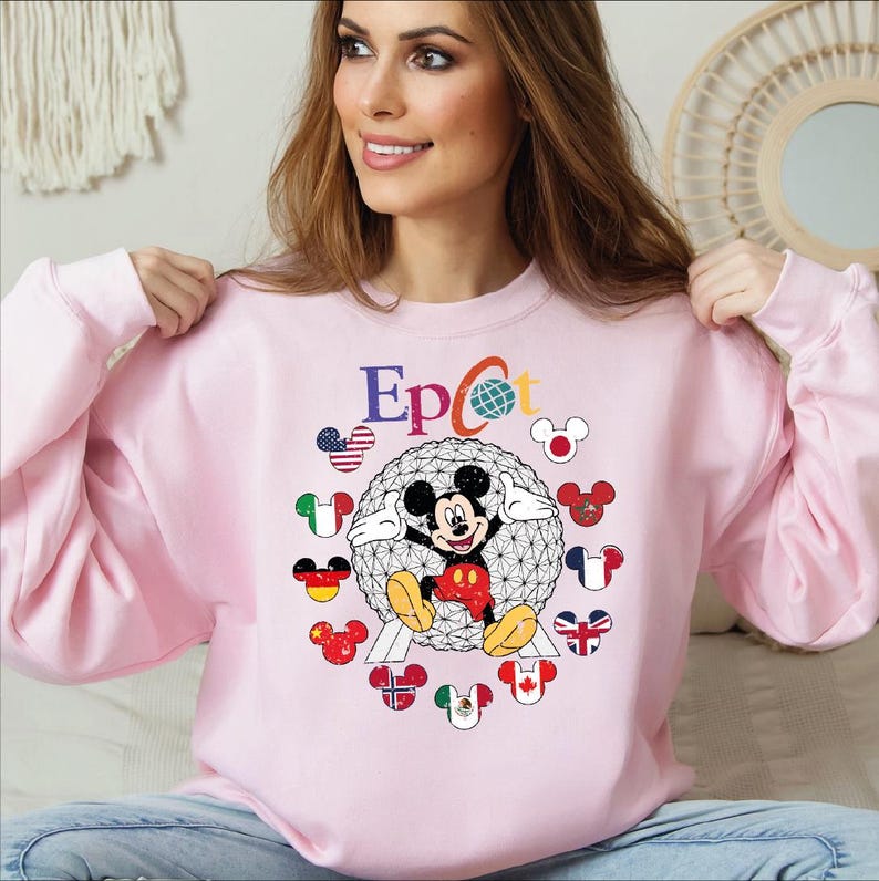 May include: A light pink sweatshirt featuring a graphic of Mickey Mouse inside a geodesic sphere, with the word "Epcot" above. Mickey is surrounded by Mickey Mouse head icons with flags. The sweatshirt has long sleeves.