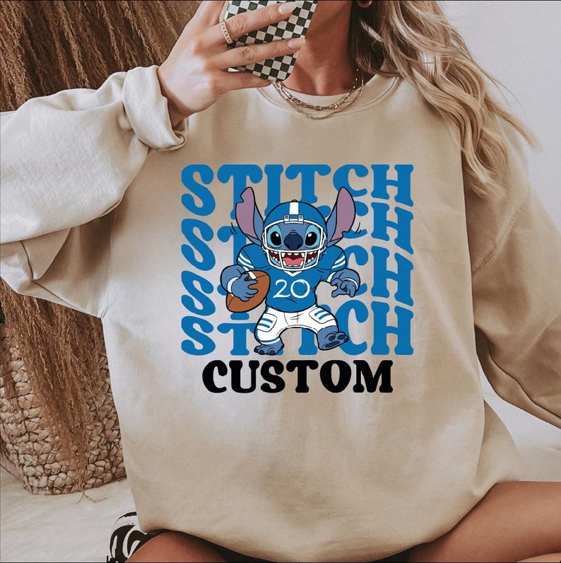 May include: Beige sweatshirt featuring a blue Stitch character in a football uniform, holding a football. The word "STITCH" is repeated in blue, with "CUSTOM" below. The character wears the number 20.