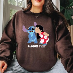 Pode incluir: Camisola castanha com um desenho de uma personagem de desenho animado azul a ser beijada por uma menina com um vestido vermelho. As palavras "CUSTOM TEXT" est&atilde;o impressas em branco. A camisola &eacute; de cor castanha lisa.