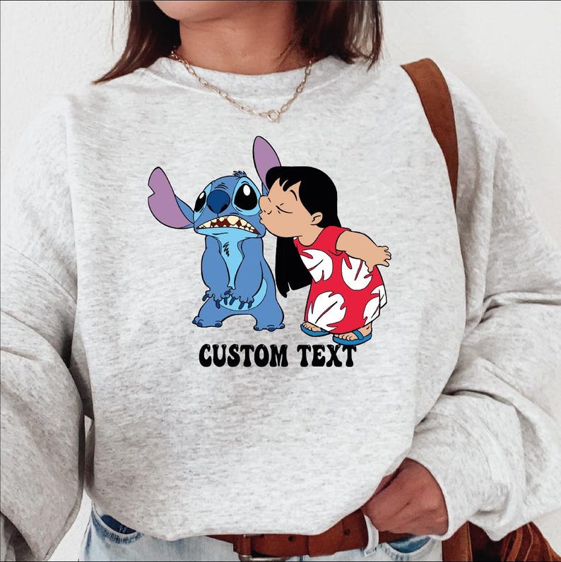 Pode incluir: Um moletom cinza claro com uma imagem de desenho animado de Stitch e Lilo do filme da Disney Lilo & Stitch. Lilo beija Stitch na bochecha. As palavras "CUSTOM TEXT" s&atilde;o impressas em preto abaixo.