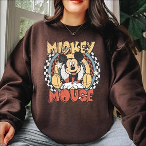 K&ouml;nnte beinhalten: Ein braunes Sweatshirt mit einem Retro-Mickey-Mouse-Motiv. Das Design zeigt die Worte "Mickey Mouse" in einer Vintage-Schrift, mit Mickey in der Mitte. Die Grafik ist vor einem Hintergrund aus einem Schachbrettmuster und Sternen platziert.