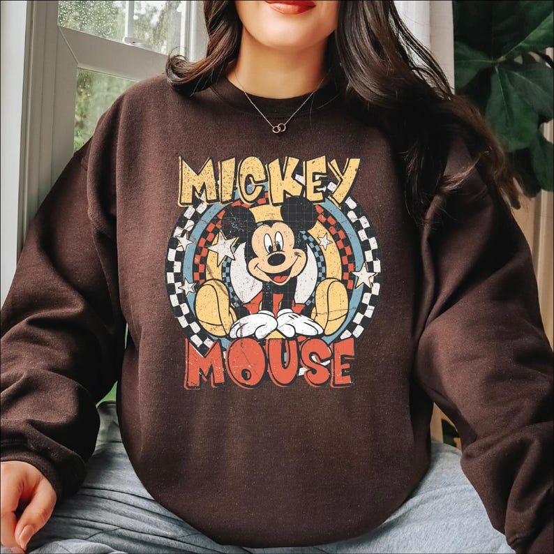 K&ouml;nnte beinhalten: Ein braunes Sweatshirt mit einem Retro-Mickey-Mouse-Motiv. Das Design zeigt die Worte "Mickey Mouse" in einer Vintage-Schrift, mit Mickey in der Mitte. Die Grafik ist vor einem Hintergrund aus einem Schachbrettmuster und Sternen platziert.