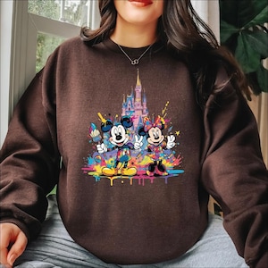 Peut inclure: Sweat-shirt marron avec un motif color&eacute; de Mickey et Minnie Mouse avec des pinceaux et un ch&acirc;teau en arri&egrave;re-plan. Le motif comprend des &eacute;claboussures de peinture vibrantes de diff&eacute;rentes couleurs. Les personnages sont dans une pose ludique.