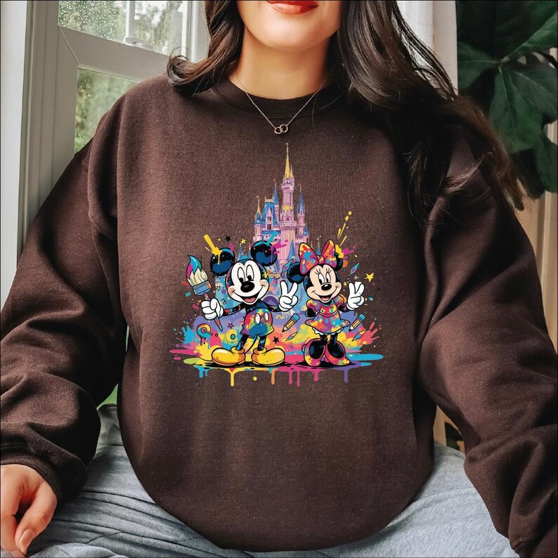 Peut inclure: Sweat-shirt marron avec un motif color&eacute; de Mickey et Minnie Mouse avec des pinceaux et un ch&acirc;teau en arri&egrave;re-plan. Le motif comprend des &eacute;claboussures de peinture vibrantes de diff&eacute;rentes couleurs. Les personnages sont dans une pose ludique.