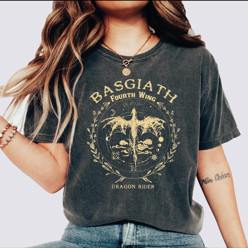 May include: Dark gray t-shirt featuring a gold dragon design within a circular frame. The text "BASGIATH FOURTH WING" arches above the dragon, with "DRAGON RIDER" below. The design includes celestial elements and floral accents.