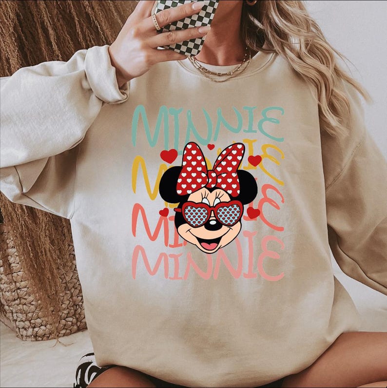 Puede incluir: Sudadera beige con un dise&ntilde;o de Minnie Mouse. El dise&ntilde;o incluye la palabra "MINNIE" en varios colores, la cara de Minnie con gafas de sol en forma de coraz&oacute;n y un lazo rojo con lunares blancos. Peque&ntilde;os corazones rojos est&aacute;n dispersos alrededor del dise&ntilde;o.