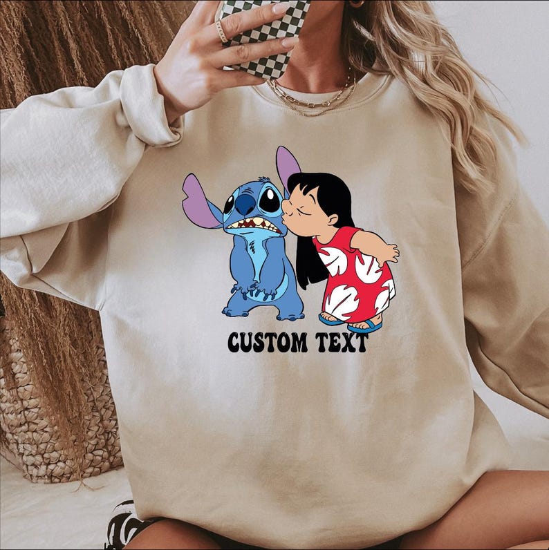 May include: A beige sweatshirt featuring a graphic of the characters Stitch and Lilo from the animated film Lilo & Stitch. Stitch is blue, and Lilo is wearing a red dress with white accents. The text "CUSTOM TEXT" is printed below the characters.