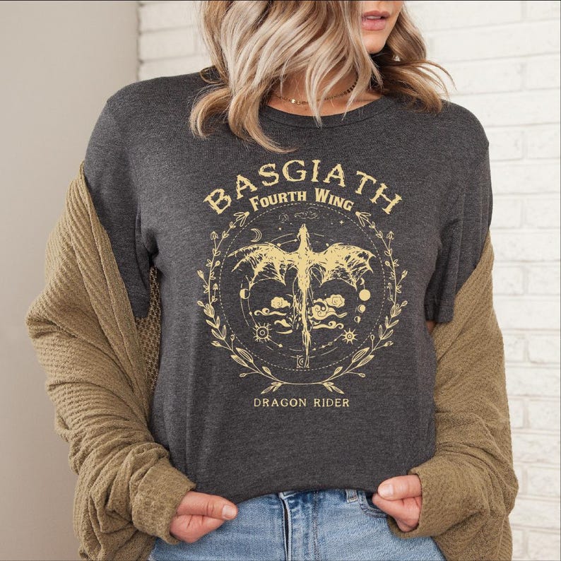 May include: Dark gray t-shirt with a gold graphic featuring a dragon, the text "BASGIATH Fourth Wing," and "DRAGON RIDER." The design is encircled by a wreath of leaves and celestial elements. The shirt has a crew neck.